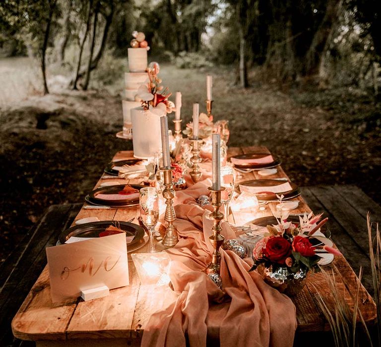 Outdoor wooden wedding tablescape with pink table runner, silver candles, white wedding cakes and tropical floral decor at festival themed wedding