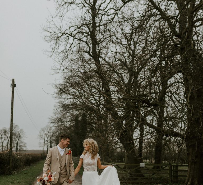 Bride with curly wedding hair wearing a two piece bridal outfit walking in the countryside with groom in relaxed beige suit