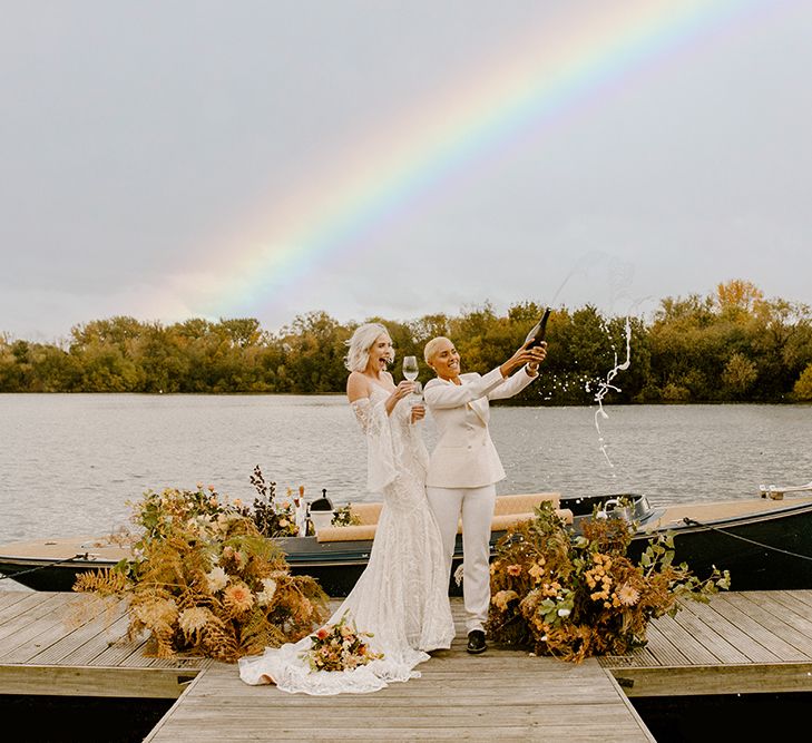 A same sex couple stands on a jeti and pops a bottle of champagne. There is a rainbow in the sky. Photography by Leah Marie Photography.