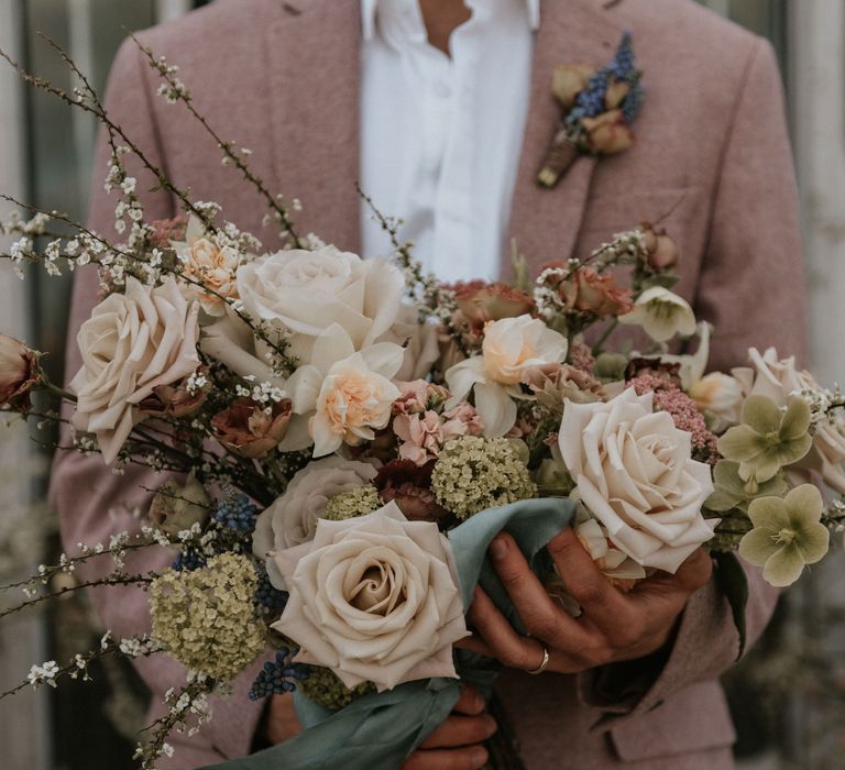 Pastel bouquet with white roses and green foliage held by groom wearing pastel pink suit 