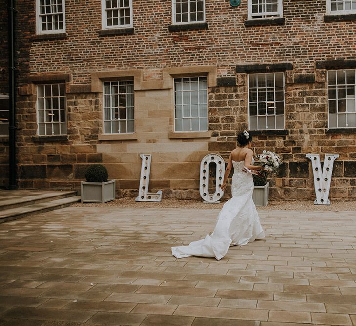 A bride wearing Sophia Tolli walks outside The West Mill wedding venue in front of large light up L O V E letters.