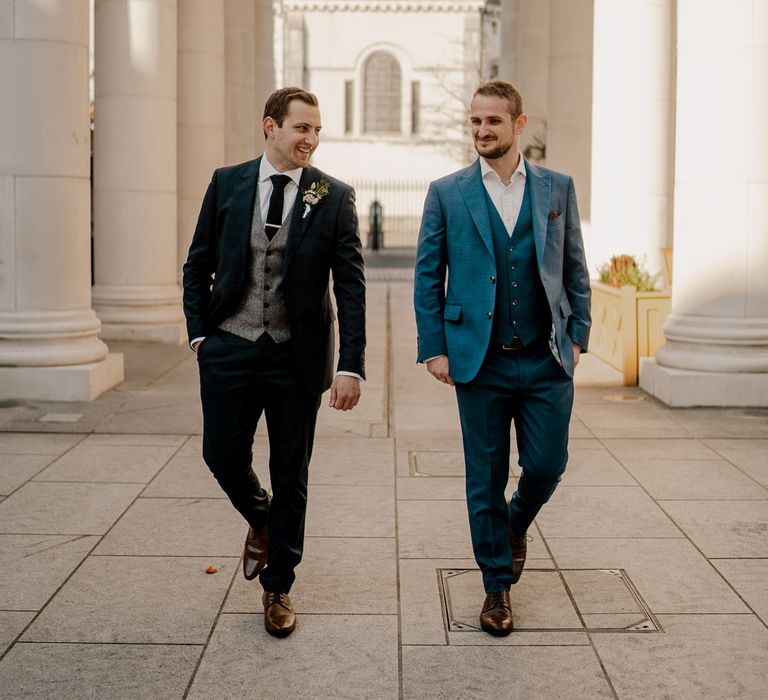 Groom in dark blue suit and grey waistcoat walks with guest in blue suit and waistcoat along paved streets before Dunluce Castle wedding