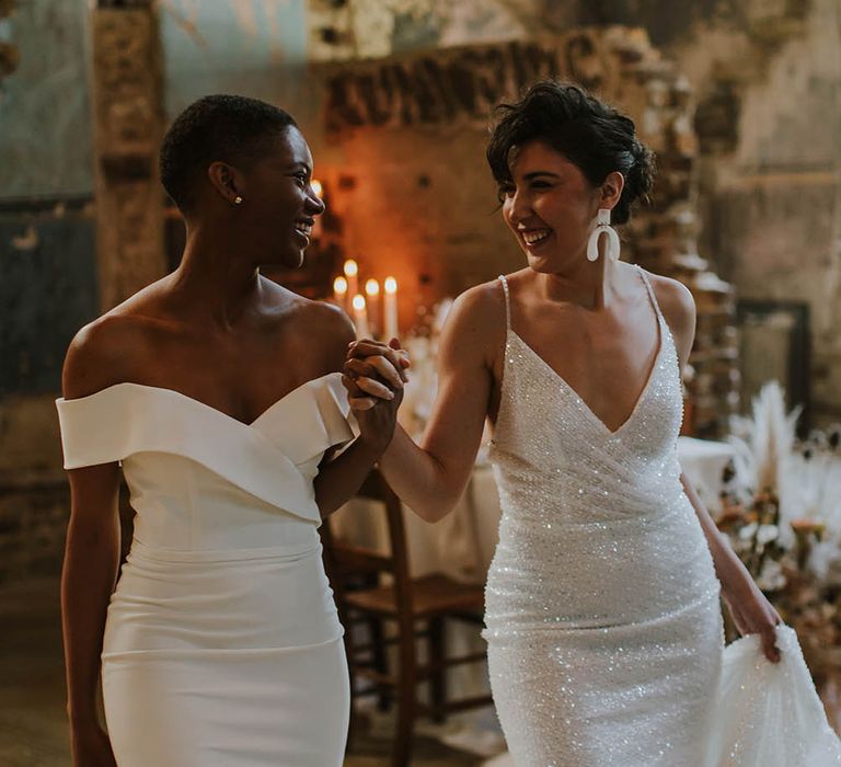 Black bride with short hair in an off the shoulder wedding dress holding hands with her bride in a sparkly wedding dress with thin straps 