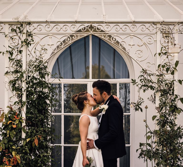 Bride & groom kiss one another outside window surrounded by green foliage