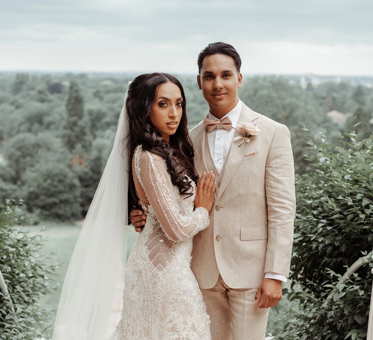 Bride & groom stand together and look toward the camera as bride wears Milla Nova wedding gown and groom wears neutral coloured suit