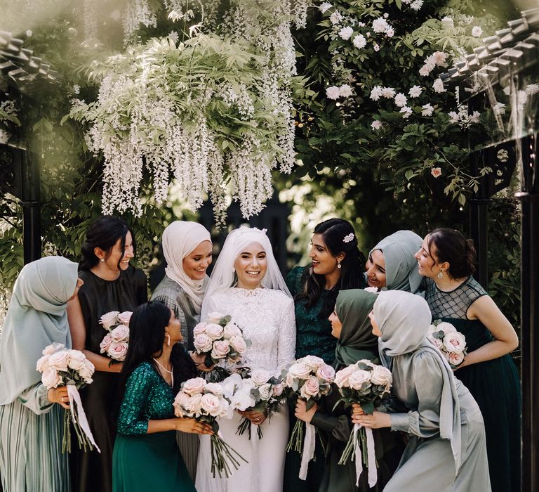 Bridesmaids smiling at the bride in various shades of green and styles of bridesmaid dress under hanging wisteria