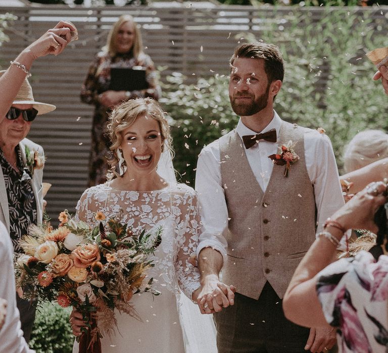 Bride in white Rime Arodaky wedding dress and mesh embroidered top holds burnt orange and peach bridal bouquet whilst holding hands with groom in waistcoat as they walk down the aisle under confetti after garden wedding ceremony with burnt orange wedding theme