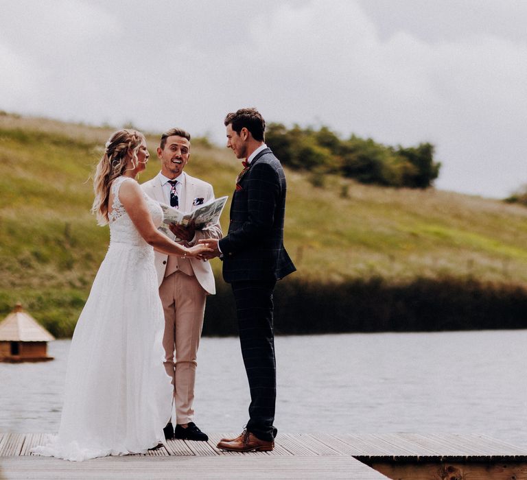 Bride & groom stand during during wedding ceremony as friend marries them in front of lake and green hills