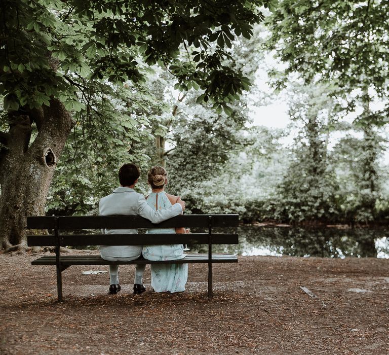 Bride & groom sit together on bench within the woodlands on the day of their wedding