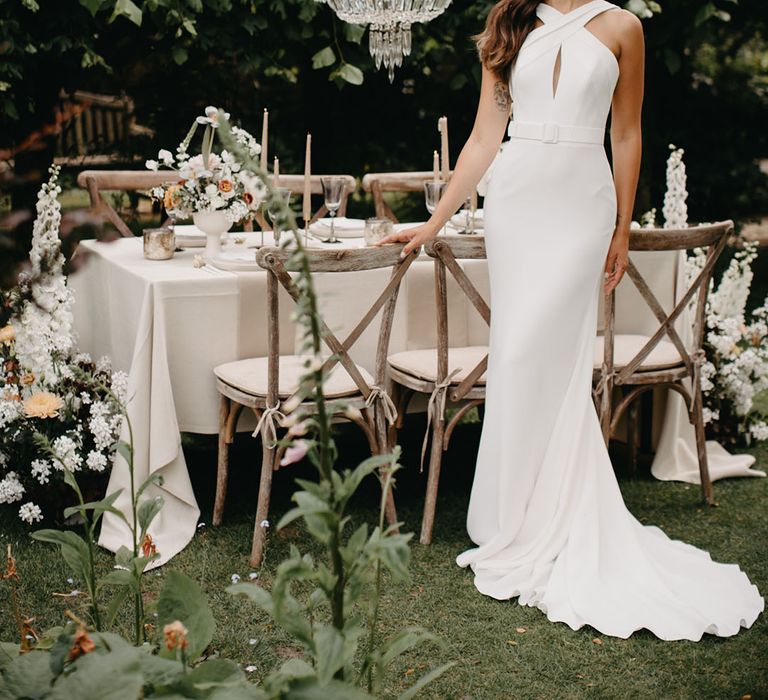 Bride in a fitted halter neck wedding dress with belt detail standing next to an outdoor table scape with chandelier installation