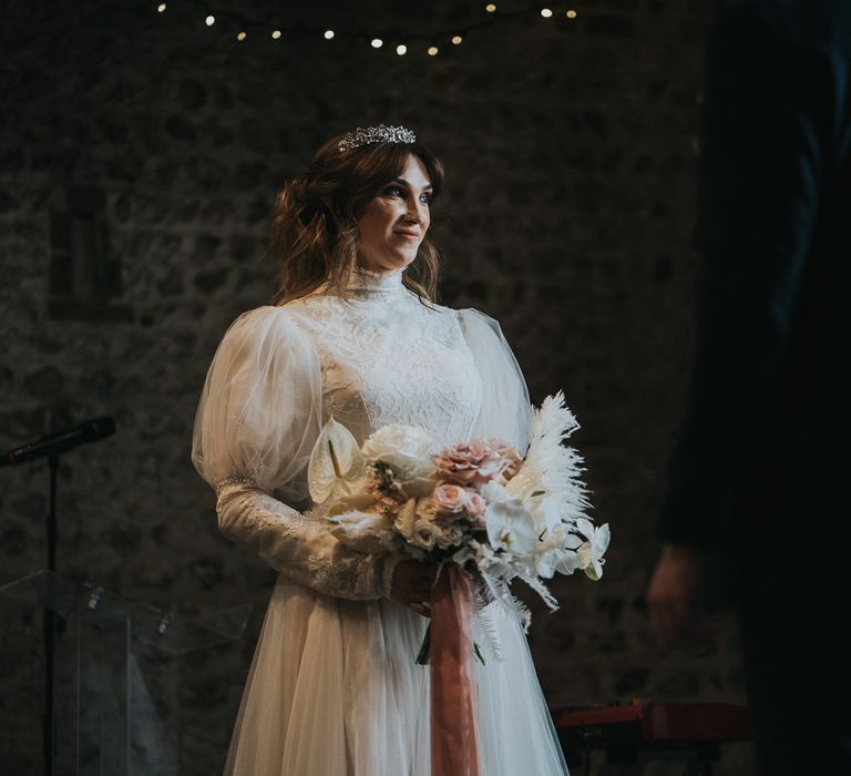 Bride holds floral bouquet tied with pink ribbon whilst wearing a high neck wedding gown with vintage gothic vibe