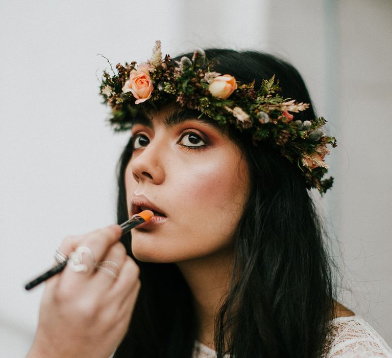 Bride with long dark hair wearing a flower crown and orange eye shadow having her beige lipstick applied. 