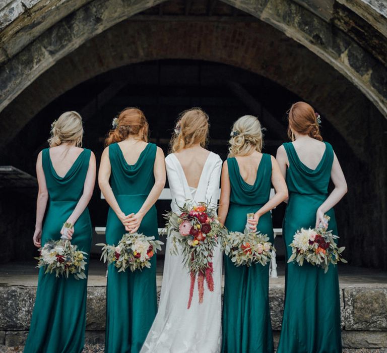 Bridesmaids in Ghost dark green satin bridesmaid dresses with cowl back detail holding their bouquets behind their backs 