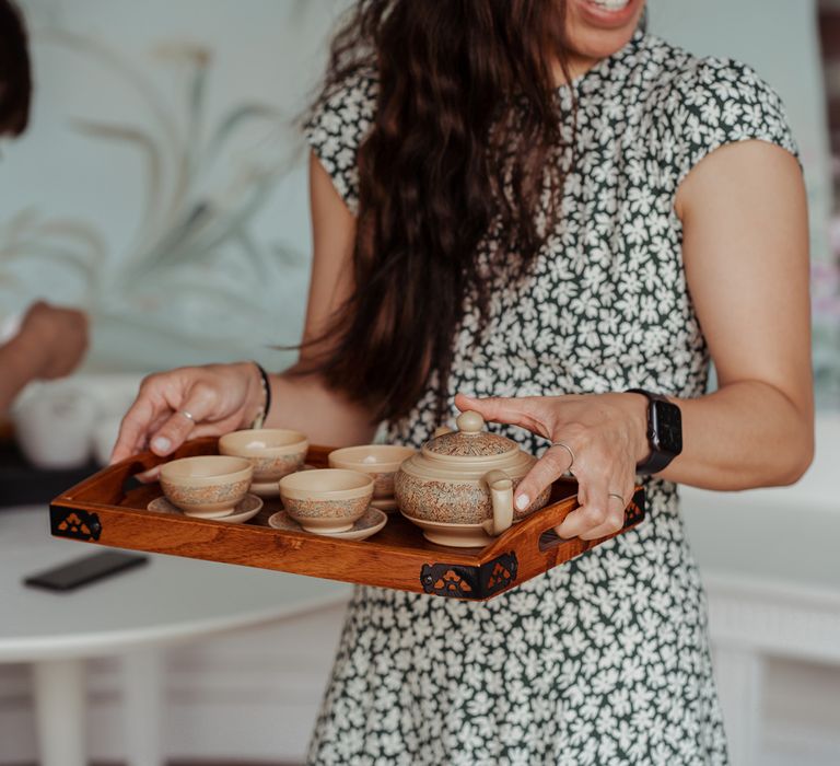 Wedding guest in black and white floral dress holds wooden tray with traditional Chinese tea set on for Chinese tea ceremony at Wasing Park wedding