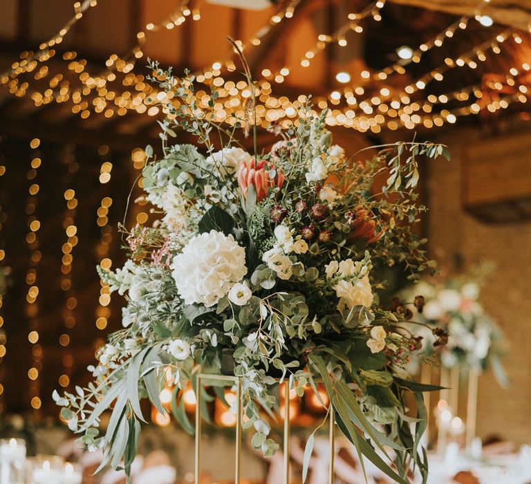 White, pink and green flower bomb on stand at long wedding breakfast table lit with strung fairy lights at Tythe Barn wedding with barn wedding flowers