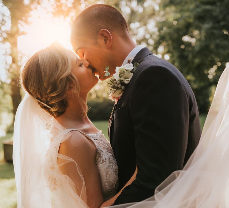 Bride in white lace Justin Alexander wedding dress and veil kisses groom in dark suit with floral buttonhole in the gardens at Tythe Barn wedding with barn wedding flowers