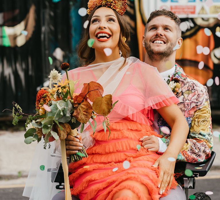 Confetti moment with bride in a coral wedding dress, Frida Kahlo flower crown and red lipstick smiling as she sits on her grooms lap in his wheelchair 