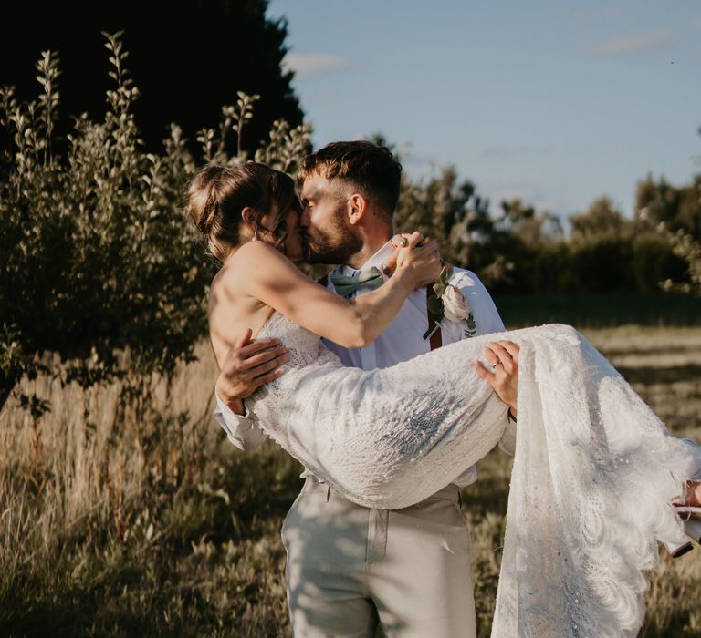 Groom carries his bride as they kiss and walk across field | Mark Bamforth Photography