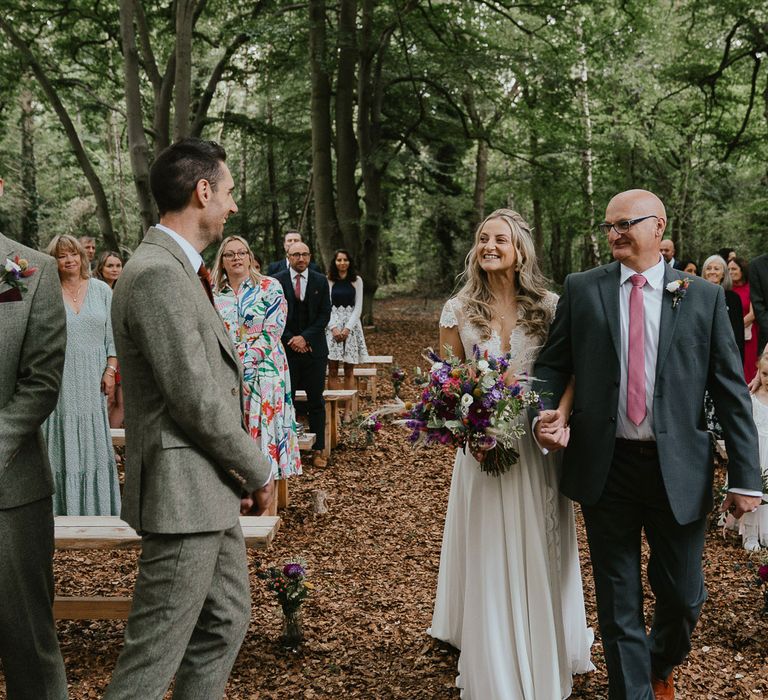 Bride in lace top wedding dress holding mixed bridal bouquet walks holding hands with father of the bride in dark suit and pink tie at woodland wedding ceremony in Norfolk