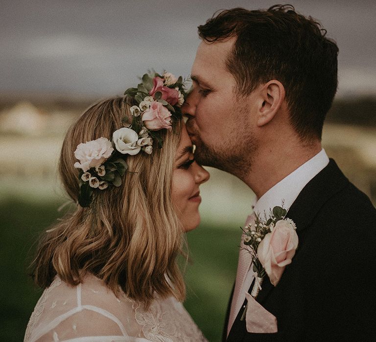 Groom in a navy blazer with a pink rose buttonhole kissing his brides forehead in a pink and white flower crown 