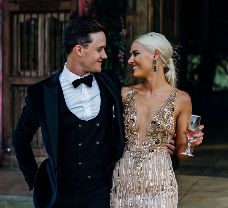 White groom in black tie and white blonde bride in gold sequin wedding dress smile at each other