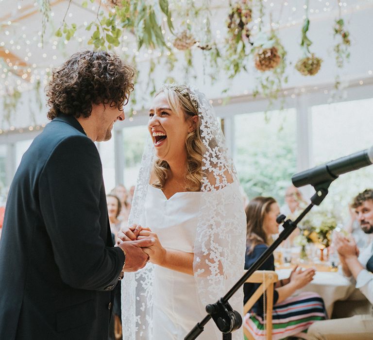 Bride & groom smile and look lovingly at one another as bride wears subtle lace embellished veil