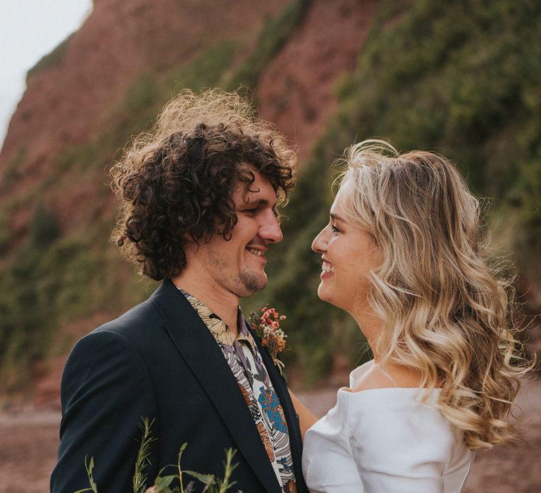 Bride & groom look lovingly at one another as they stand on the beach