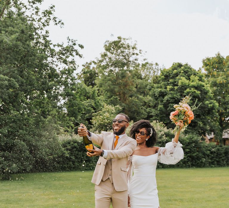 Black bride in a strapless wedding dress with sheer sleeves and front slit holding her coral peony bouquet in the air as her groom in a beige suit pops champagne 