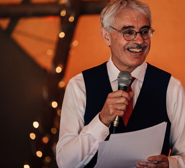 Wedding guest in white shirt, red tie and black waistcoat holds microphone as he makes speech at tipi wedding reception 