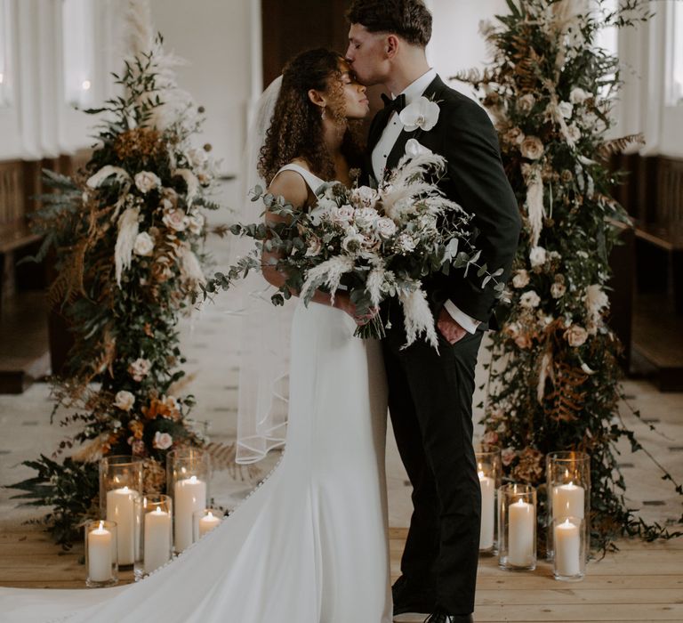 Groom kisses the top of his brides head as he wears black tie complete with white floral buttonhole 