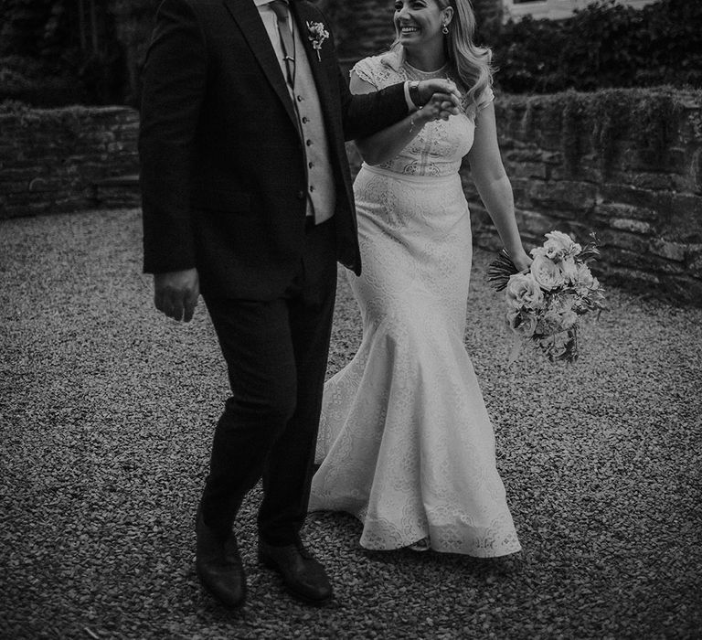 Black and white picture of the bride in a lace wedding dress and groom in a three-piece suit holding hands at their Dewsall Court wedding 