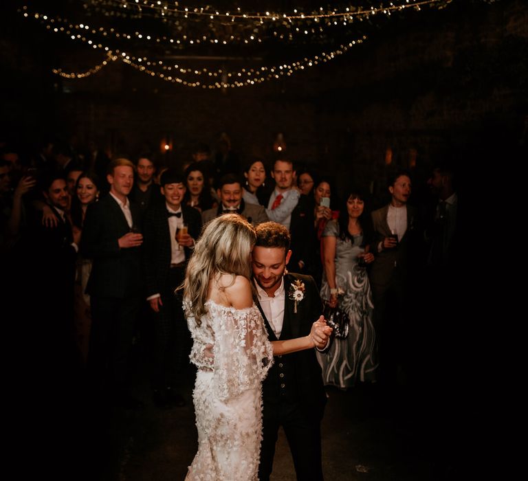 Guests watch as bride in boho bardot wedding dress dances with groom in dark suit and buttonhole under fairy light canopy at rustic wedding reception