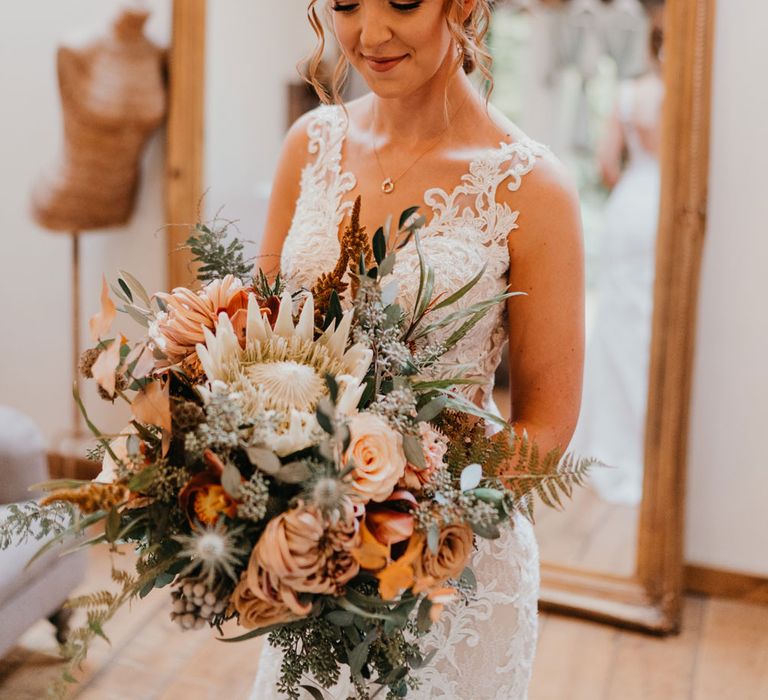 Bride with braided updo holding large mixed bridal bouquet and lace wedding dress stands in bedroom before wedding ceremony