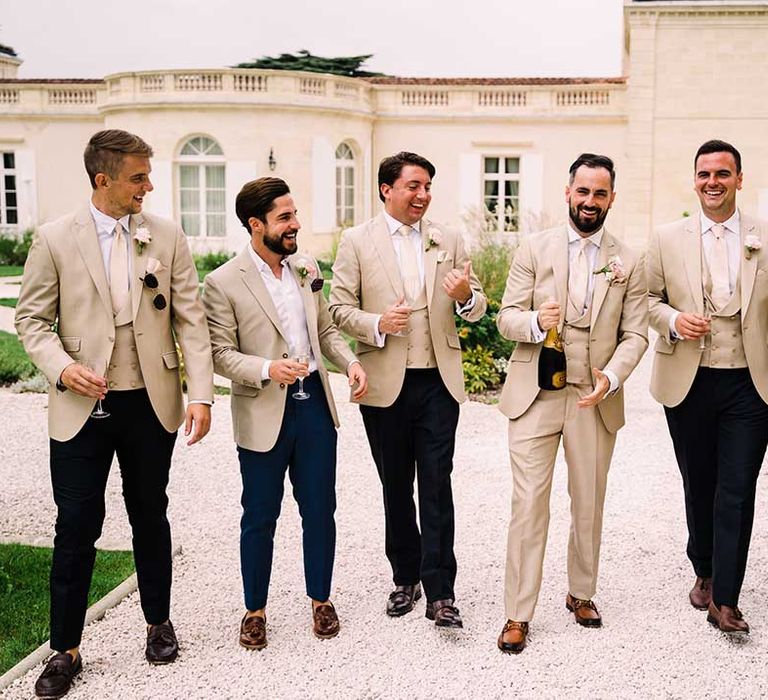 Groom and his groomsmen wearing beige suits stand outside chateau on his wedding day and pop champagne 