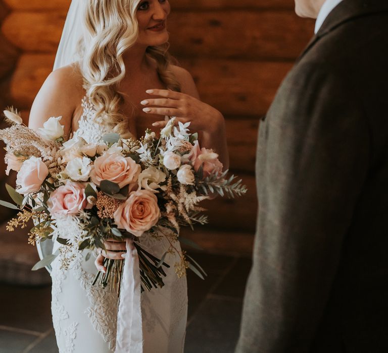 Bride with blonde hair in lace wedding dress and veil holding mixed pink and white bridal bouquet smiles at man in suit before rustic wedding