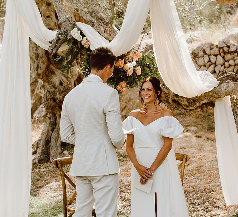 Smiling bride in Alexandra Grecco wedding dress as groom in beige suit reads wedding vows