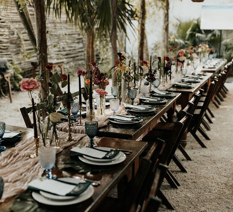 Long wooden tables with green themed tablescape with dark flowers and coloured glassware at outdoor wedding