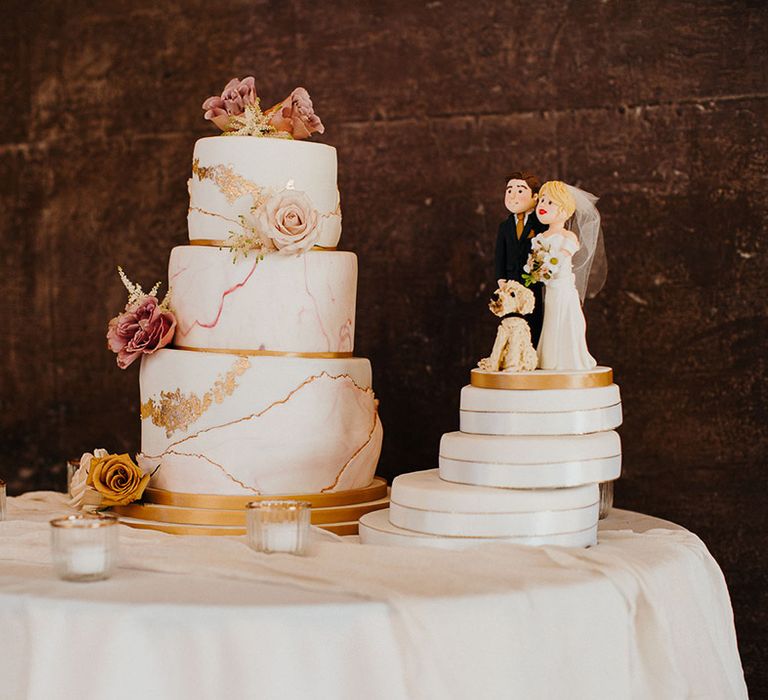 White and gold three-tier marble effect wedding cake with rose decoration next to customised cake topper of the couple and their dog