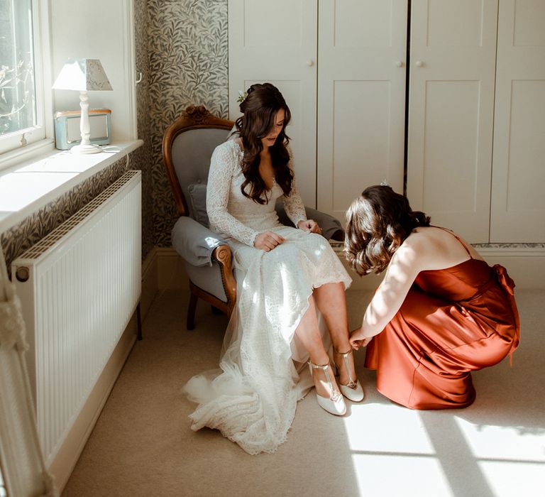 Bridesmaid in orange satin dress helps the bride put on her white sparkly wedding shoes with a rounded toe 