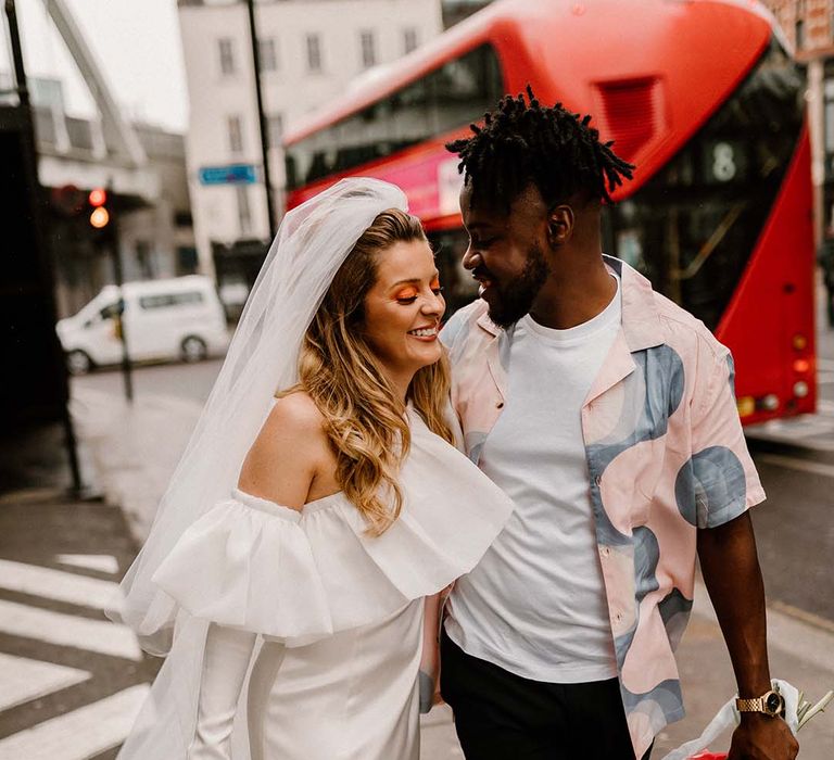 Bride in a short Bardot wedding dress with long sleeves and long veil walking through London with her groom in a patterned shirt 