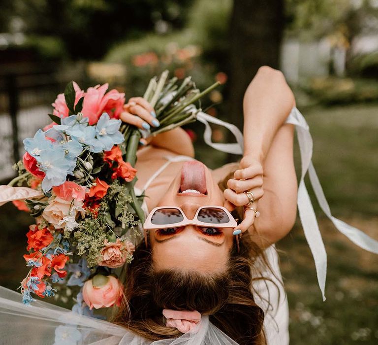 Bride leaning over in pink sunglasses holding a coral pink and blue wedding bouquet with peonies, anthuriums and delphiniums