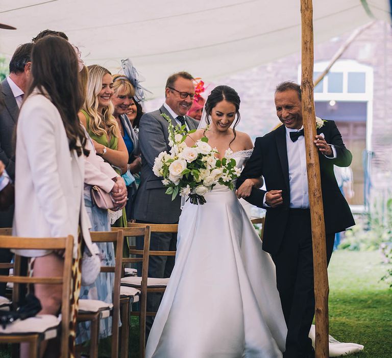 Father of the bride in black tie walks his daughter in sparkly off the shoulder v-neck wedding dress down the aisle