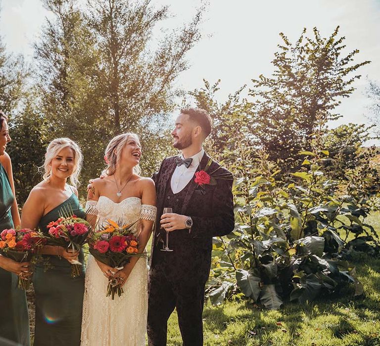 Bridesmaids with one shoulder emerald green dresses with bride and groom holding colourful bouquets and groom with red rose boutonnière