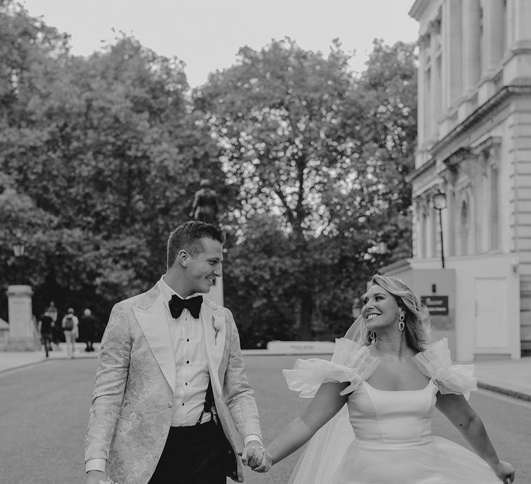 Bride and groom look at each other as they walk around London on their wedding day as groom carries the white tulip bridal bouquet