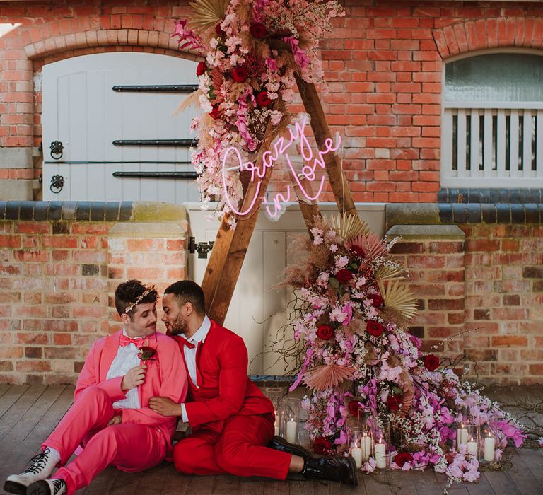 Two grooms in pink and red suits sitting in front of a wooden triangle frame altar decorated with neon sign and pink flower with dried grasses 