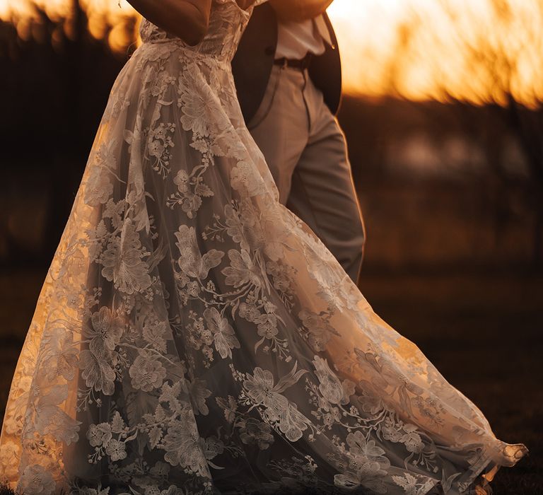 Golden hour shot of the bride and groom dancing together with bride in floral lace wedding dress and groom in grey suit jacket 