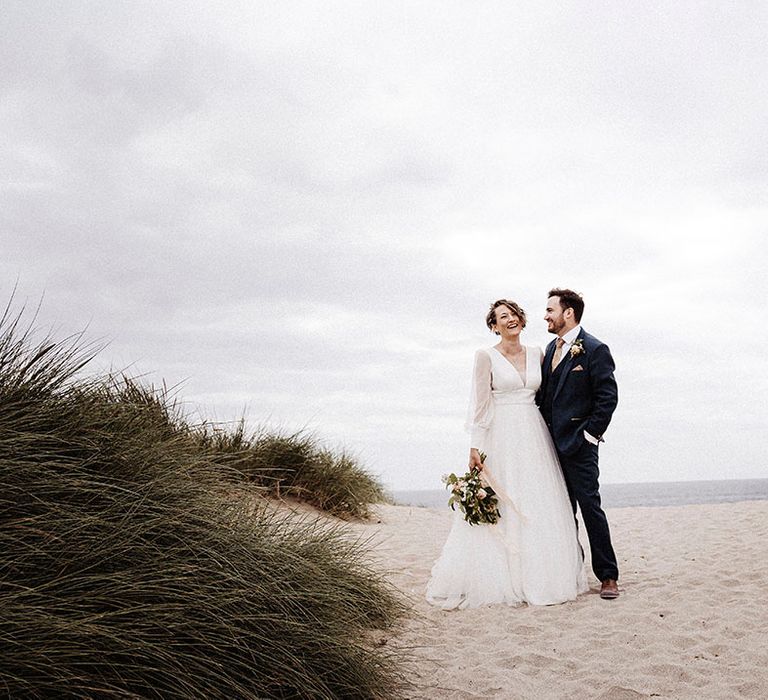 Bride and groom stand with each other on a beach in Cornwall on their wedding day with bride in tulle wedding dress and groom in blue suit