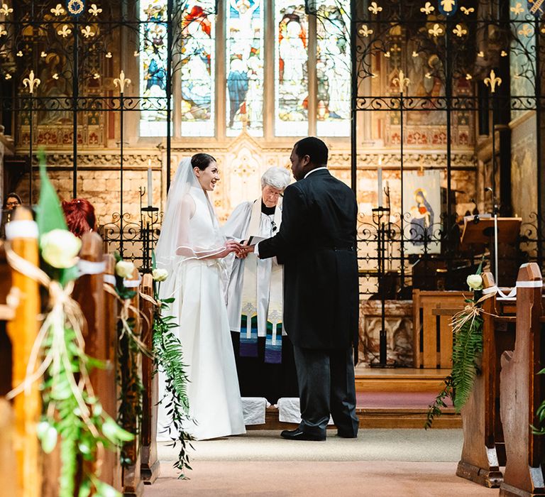 Bride in halter neck Jesus Peiro wedding dress places the ring on the groom's finger during their church wedding ceremony 