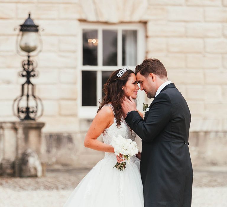 Groom in morning suit with striped trousers cups the bride's face in a princess style wedding dress and tiara crown with white rose bouquet 