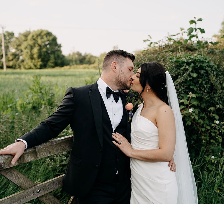 Bride and groom share a kiss as they stand in front of wooden gate at their rustic barn wedding venue 